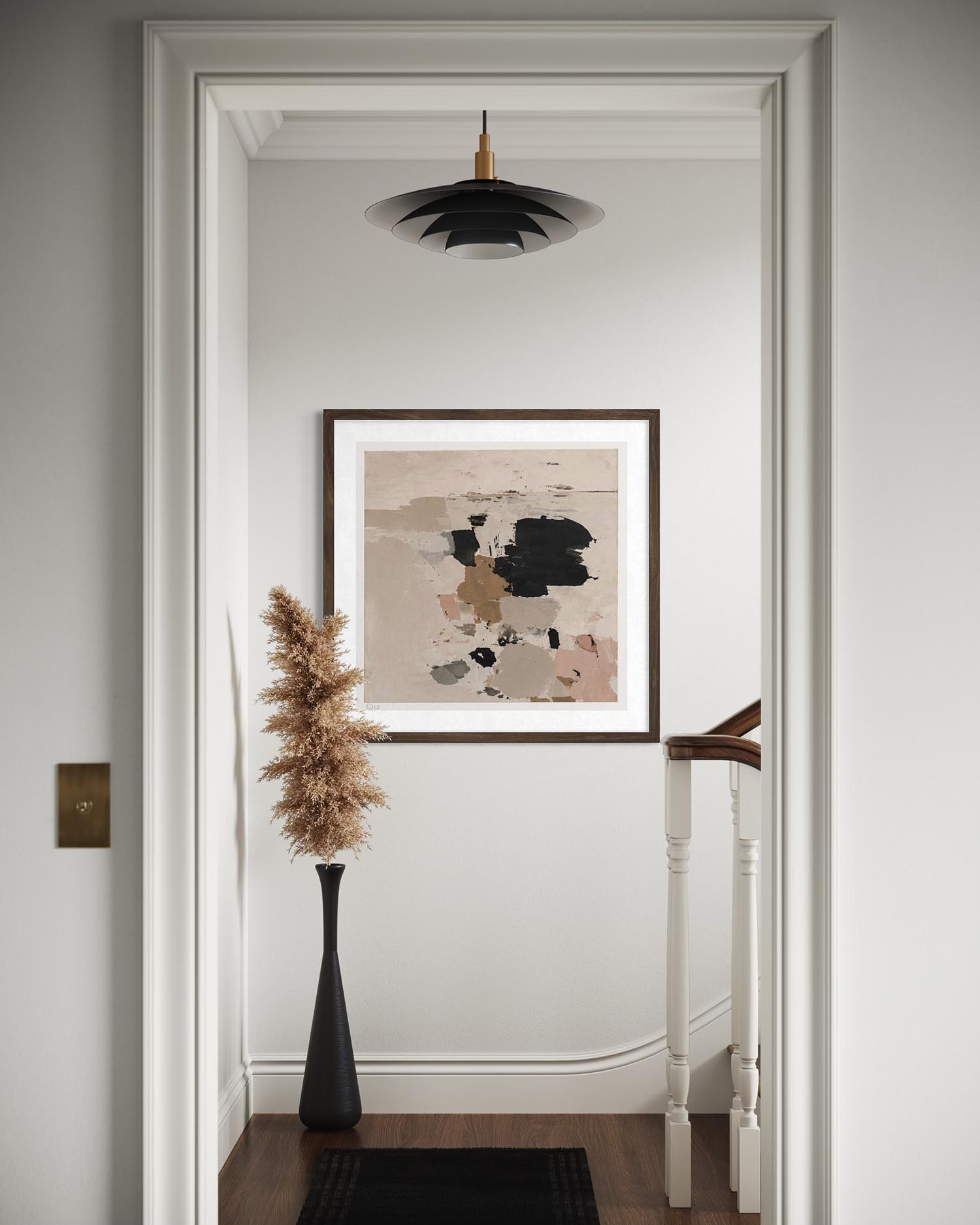 A framed abstract art print with beige, black, and brown tones hangs on a white wall in a hallway, with a black vase of dried grass and a black pendant light above.