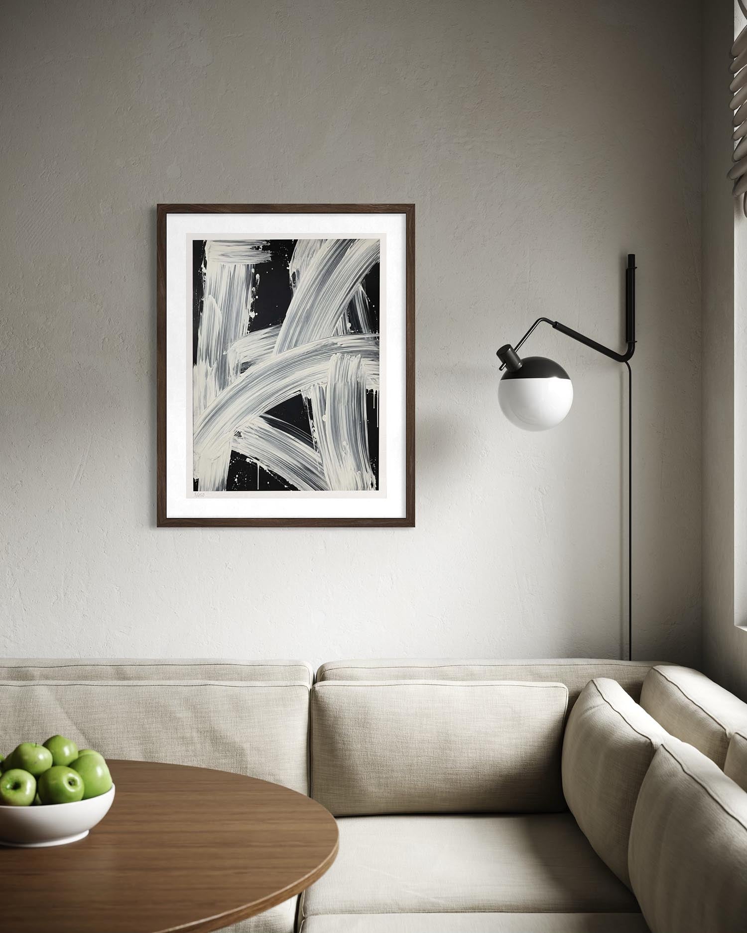 A framed abstract black and white art print hangs on a textured gray wall above a beige sectional sofa, with a wooden side table holding green apples in the foreground and a black floor lamp to the right.