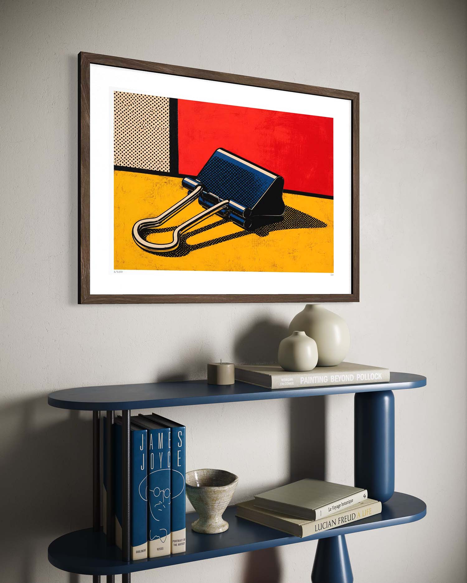 A framed pop art print featuring a black binder clip on a yellow and red background, displayed on a light gray wall above a blue shelf with books and decorative objects.