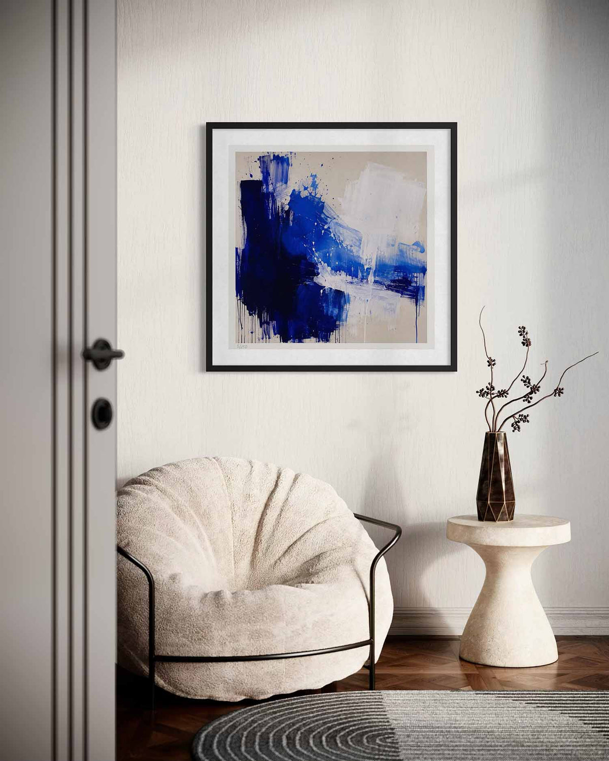 A framed abstract painting with blue, navy, white, and beige brushstrokes hangs on a light gray wall above a cream-colored lounge chair and a small side table with a vase.