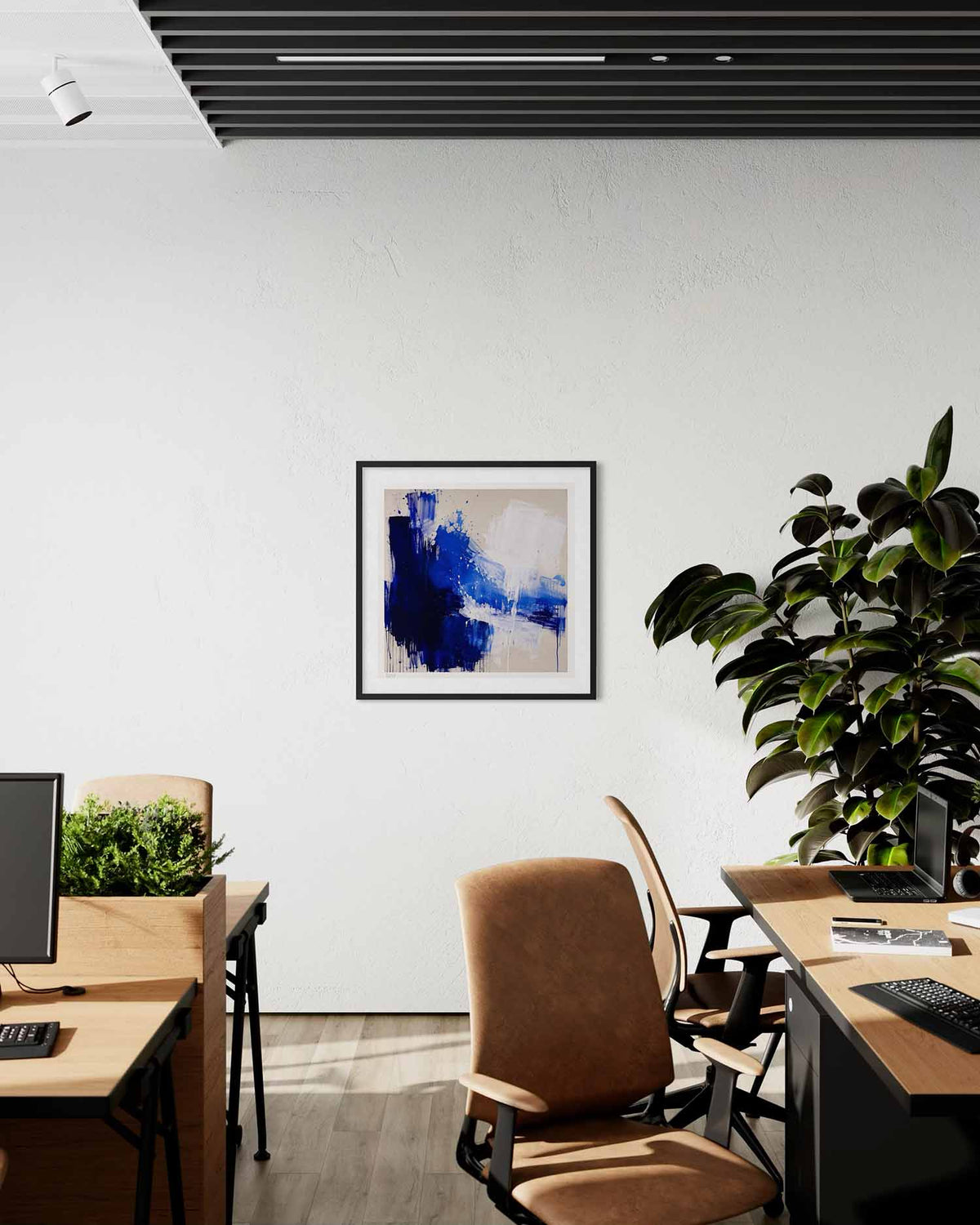 A framed abstract art print with blue, navy, and white brushstrokes hangs on a white wall in a modern office setting with desks, chairs, and a potted plant.