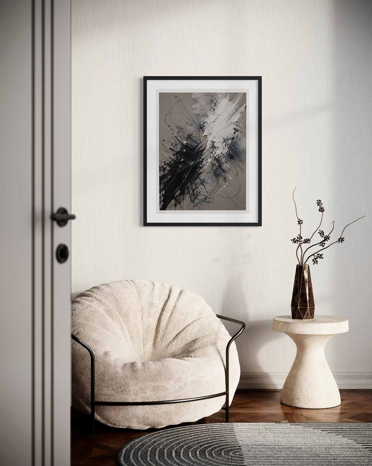A framed abstract art print with black, gray, and white brushstrokes hangs on a textured off-white wall in a modern living room setting. Below the frame is a cream-colored lounge chair with a black metal frame, and to the right is a small round side table holding a dark brown vase with dried branches.