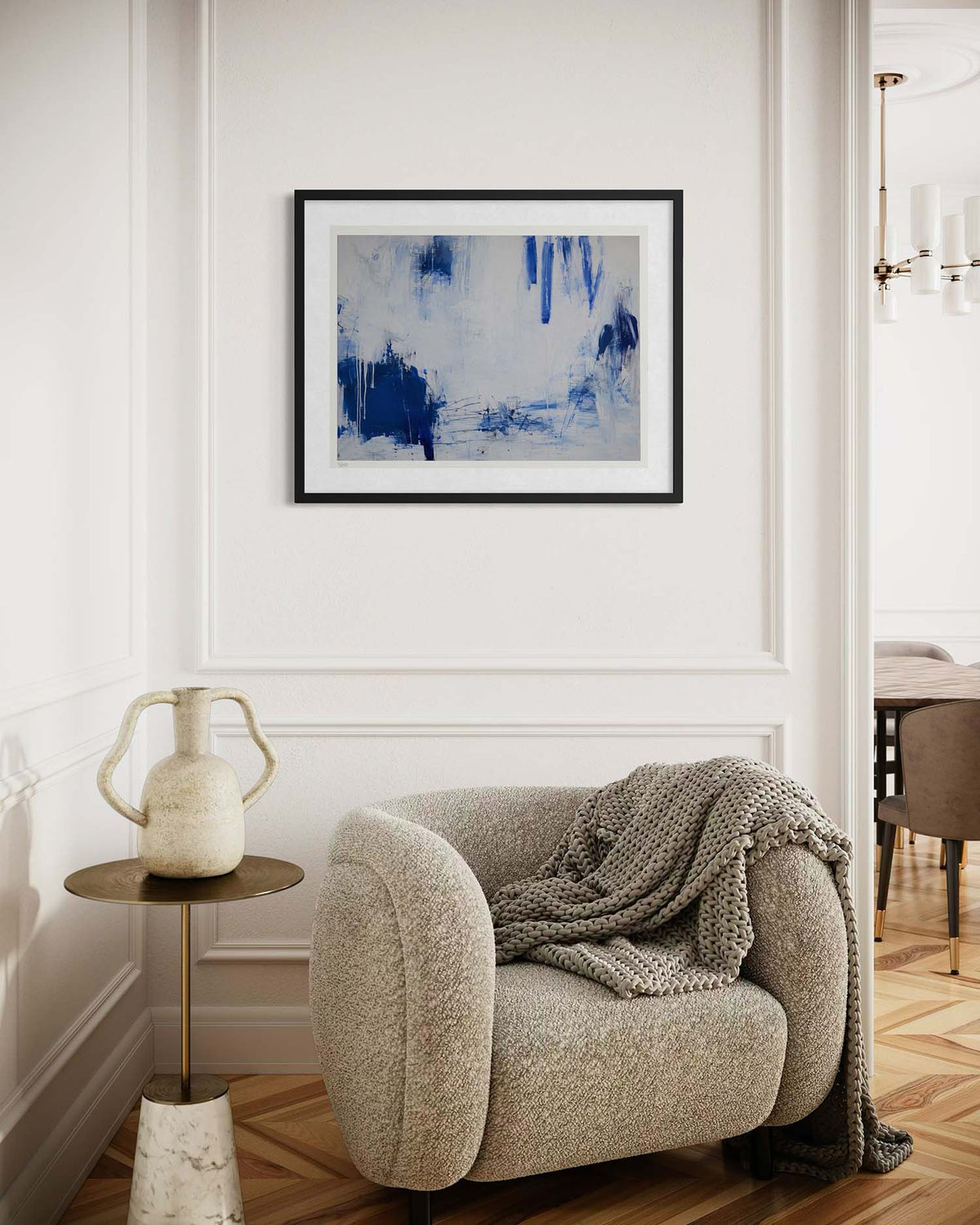 A framed abstract blue painting hangs on a white paneled wall above a beige armchair with a gray knit throw in a bright, modern living room.