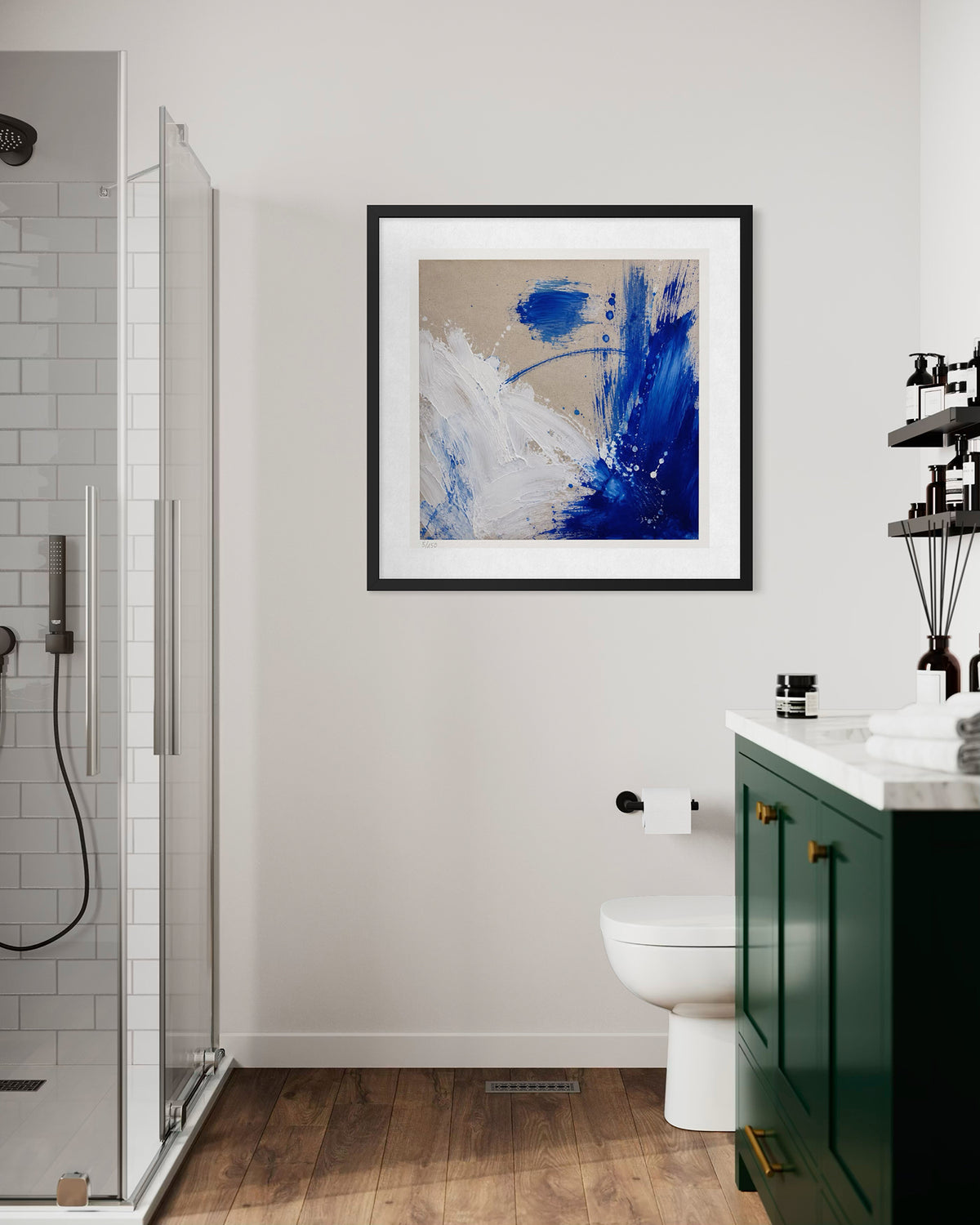 Framed abstract wall art with blue, white, and beige brushstrokes hanging on a light gray bathroom wall above a toilet, with a green vanity and wood floor visible.