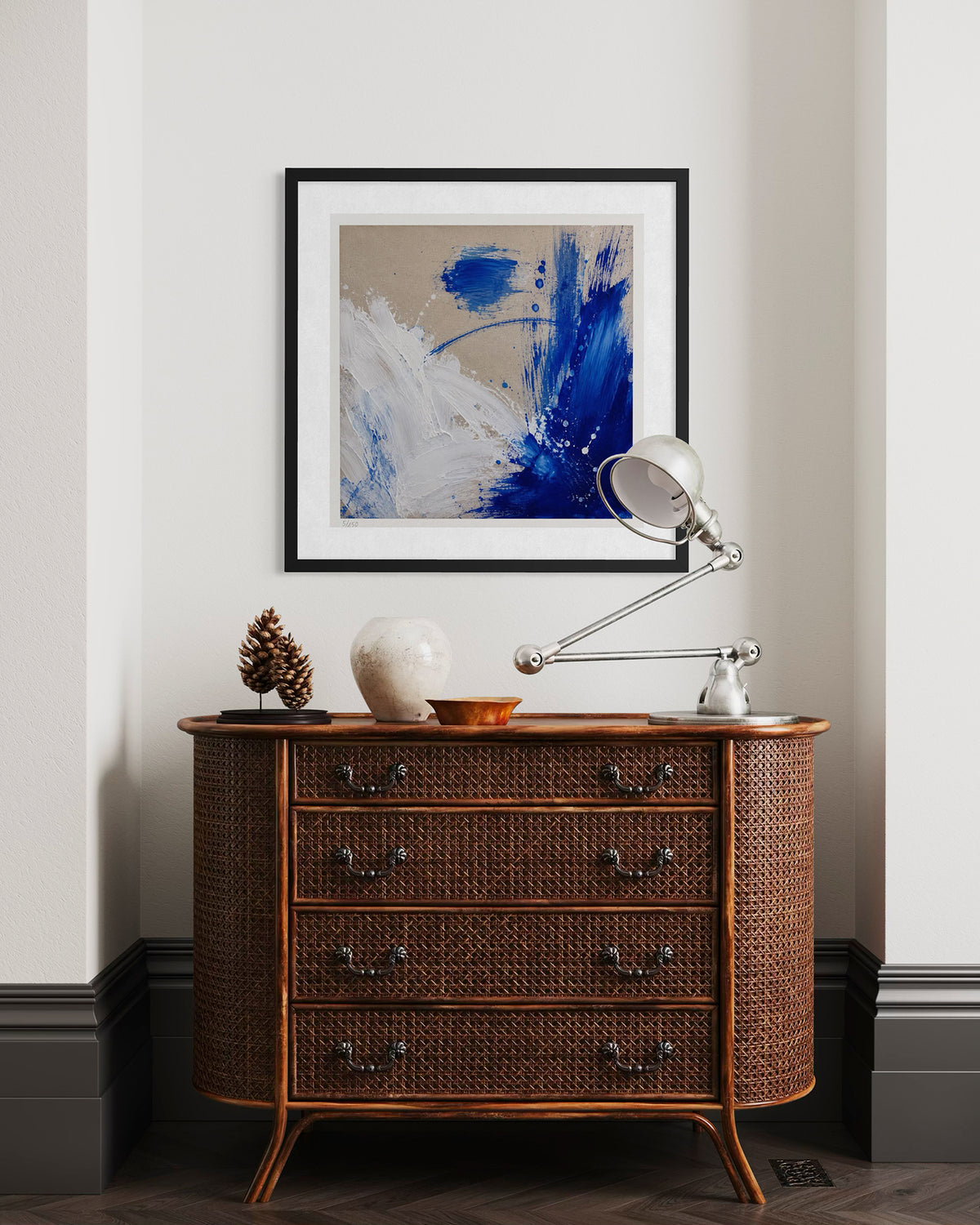 A framed abstract art print with blue, white, and beige brushstrokes hangs on a light gray wall above a brown woven wood chest of drawers. A silver desk lamp and decorative objects sit on the dresser.