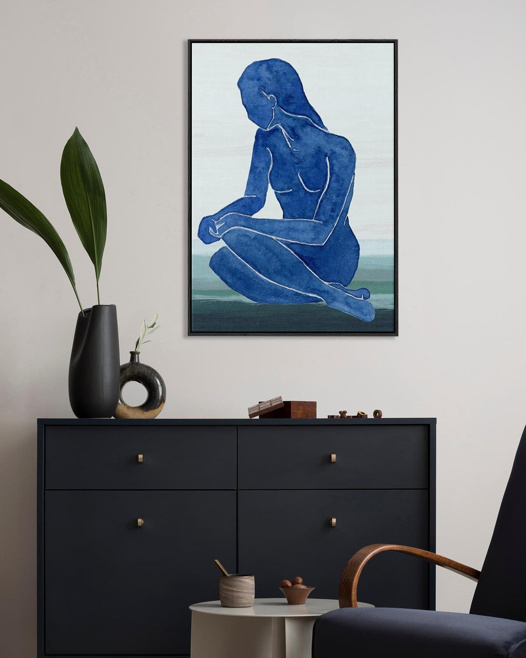 Framed abstract art of a blue figure on a wall above a black dresser.