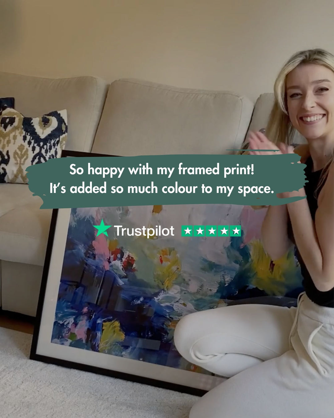 Woman sitting on a couch with a framed colorful print, smiling and holding a Trustpilot review card.