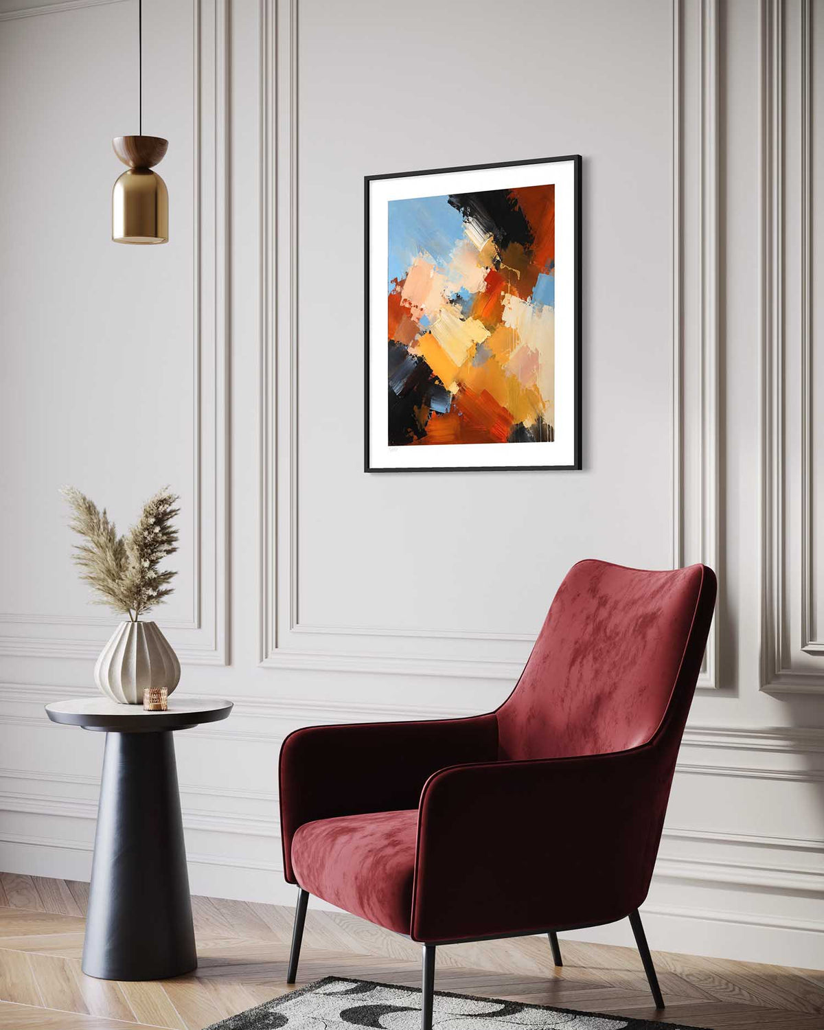 An abstract framed art print hangs on a light gray paneled wall above a dark red velvet armchair, with a small black side table and vase holding dried grass in the foreground.