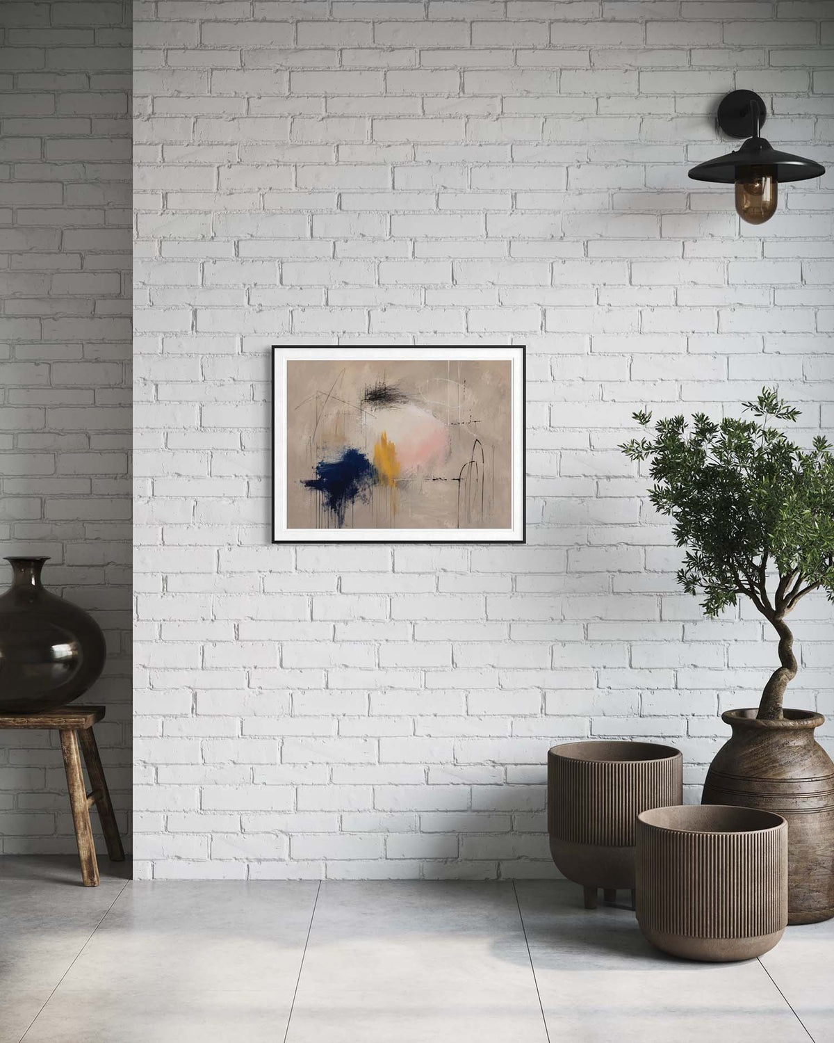 An abstract framed wall art print hangs on a white brick wall in a styled interior setting with a black pendant light, a potted tree, and ceramic vases.