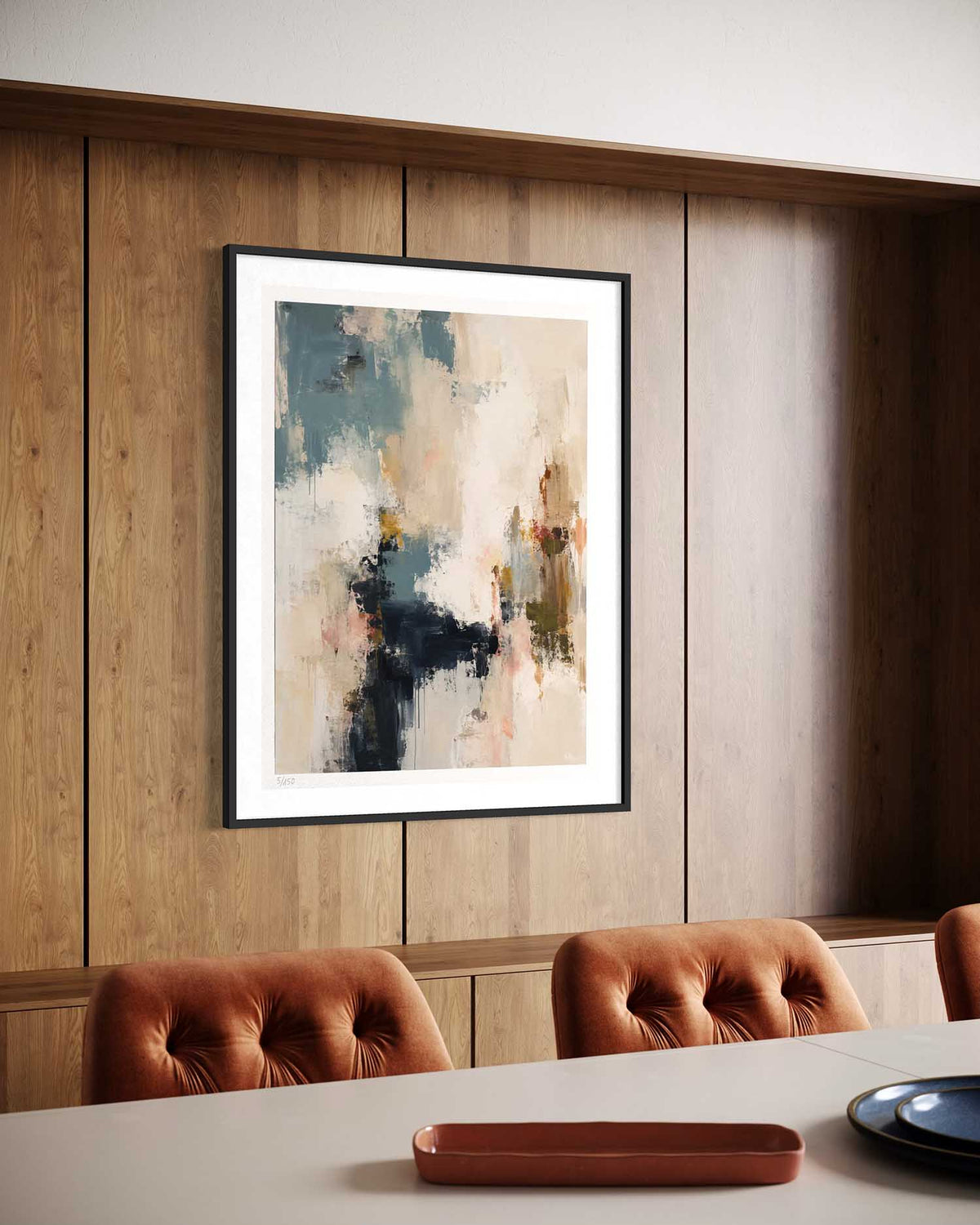 A framed abstract art print with a white mat and black frame hangs on a wood-paneled wall above a dining table with tufted brown chairs.