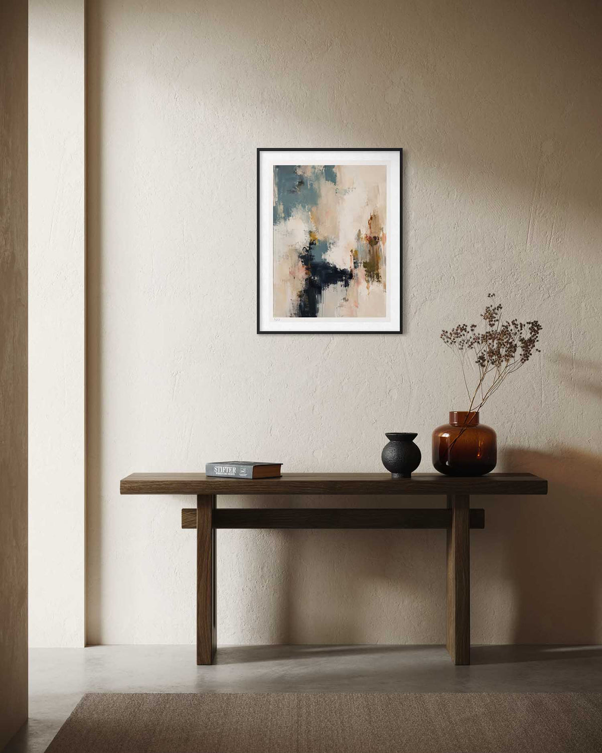A framed abstract art print with black, beige, and blue tones hangs on a textured beige wall above a dark wood console table styled with a book, a small black vase, and an amber glass vase with dried branches.