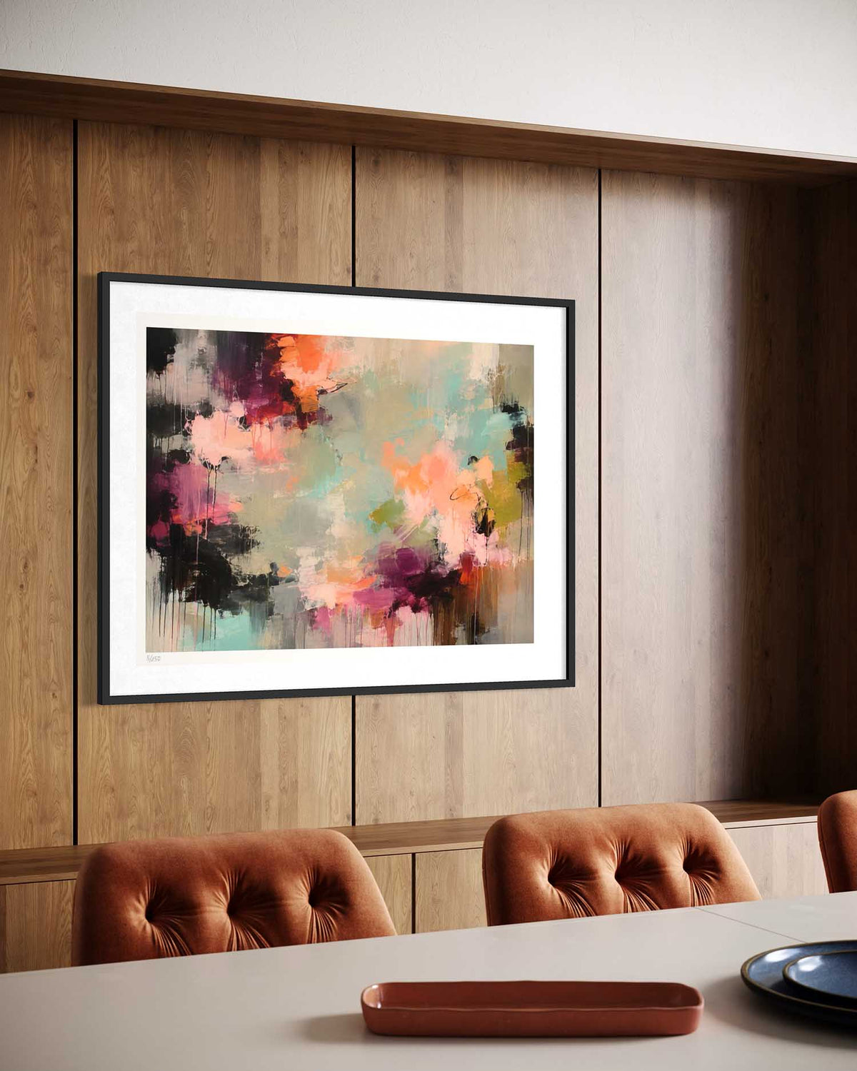 Framed abstract wall art hanging on a wood-paneled wall above a dining table with tufted chairs. The artwork features a colorful abstract composition with pink, orange, teal, purple, black, and cream tones, set in a black frame with a white mat.