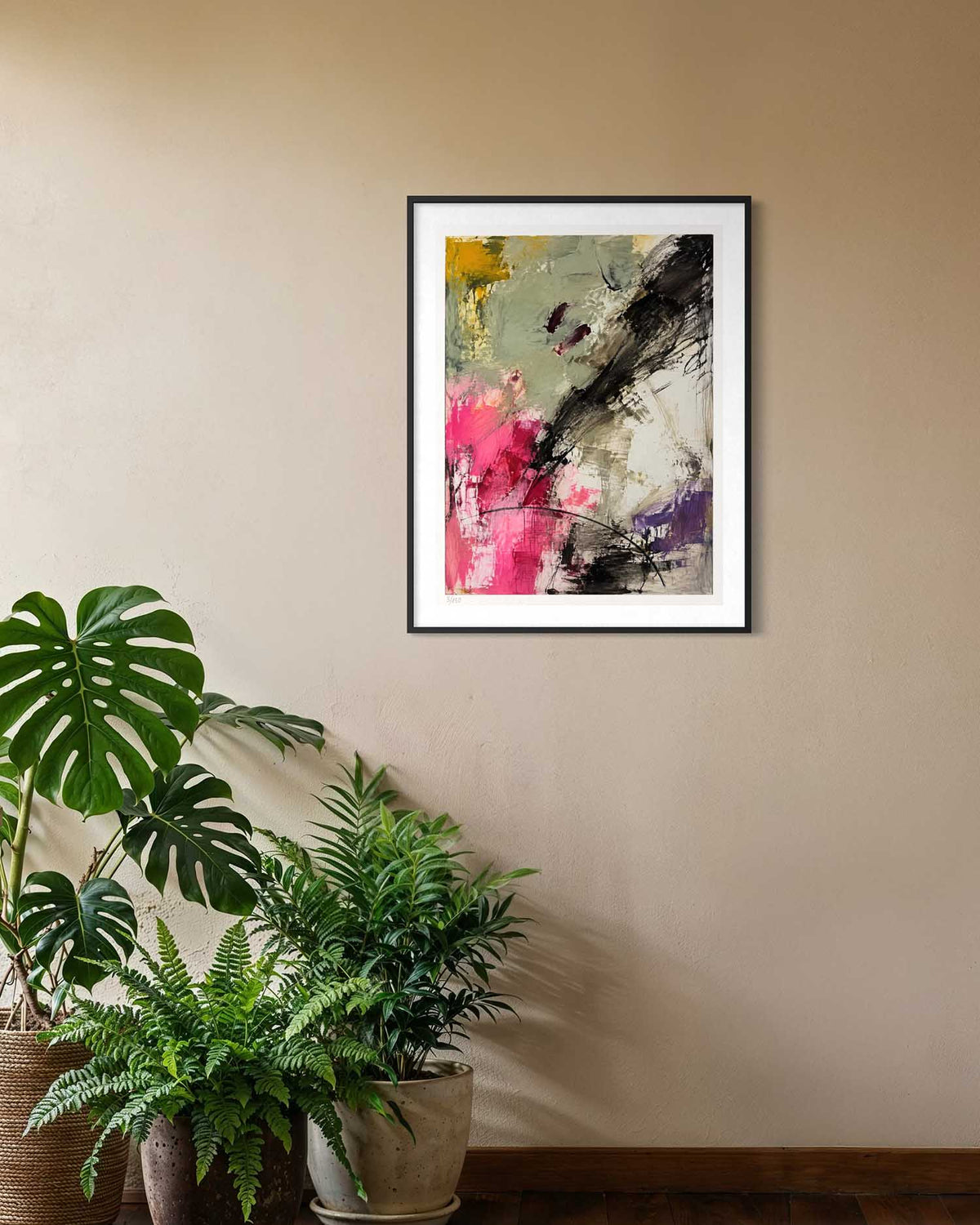 A framed abstract art print featuring vibrant pink, black, gray, and yellow brushstrokes, displayed on a beige wall above potted green plants.