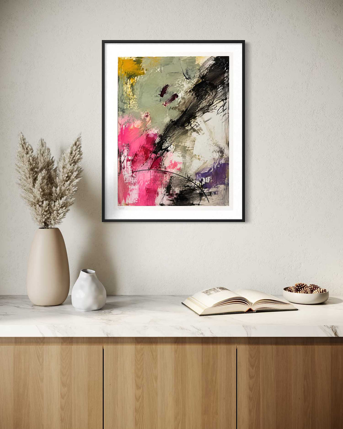 A framed abstract wall art print hangs on a light beige wall above a marble countertop with a wooden cabinet below. The artwork features expressive brushstrokes in black, gray, pink, yellow, and purple on a white mat, with a black frame.