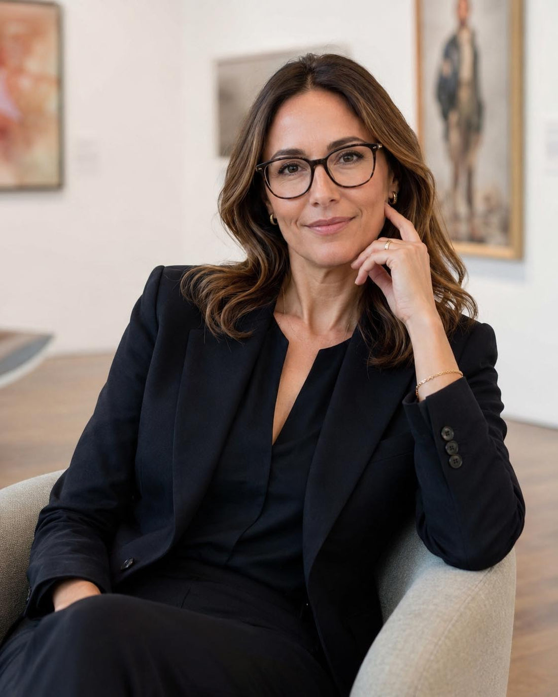 A woman with brown hair and glasses sits in a light beige armchair, wearing a black blazer and matching trousers in a bright, modern art gallery setting.