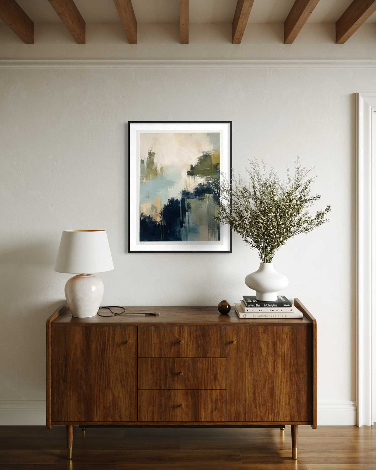 A framed abstract landscape art print hangs on a light beige wall above a mid-century modern wooden sideboard. The artwork features blue, green, and beige tones.