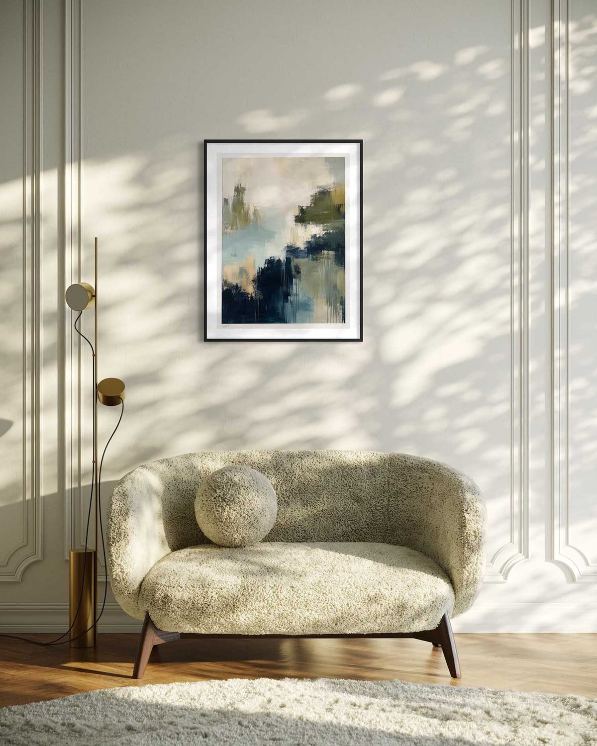 A framed abstract art print with blue, green, and beige tones hangs on a paneled wall above a textured beige armchair in a sunlit room with leaf shadows.