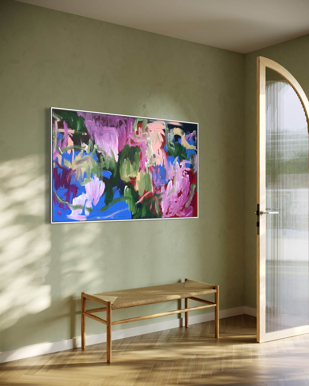 A colorful abstract canvas painting with pink, blue, green, and purple brushstrokes, displayed on a sage green wall in a modern room above a woven bench.