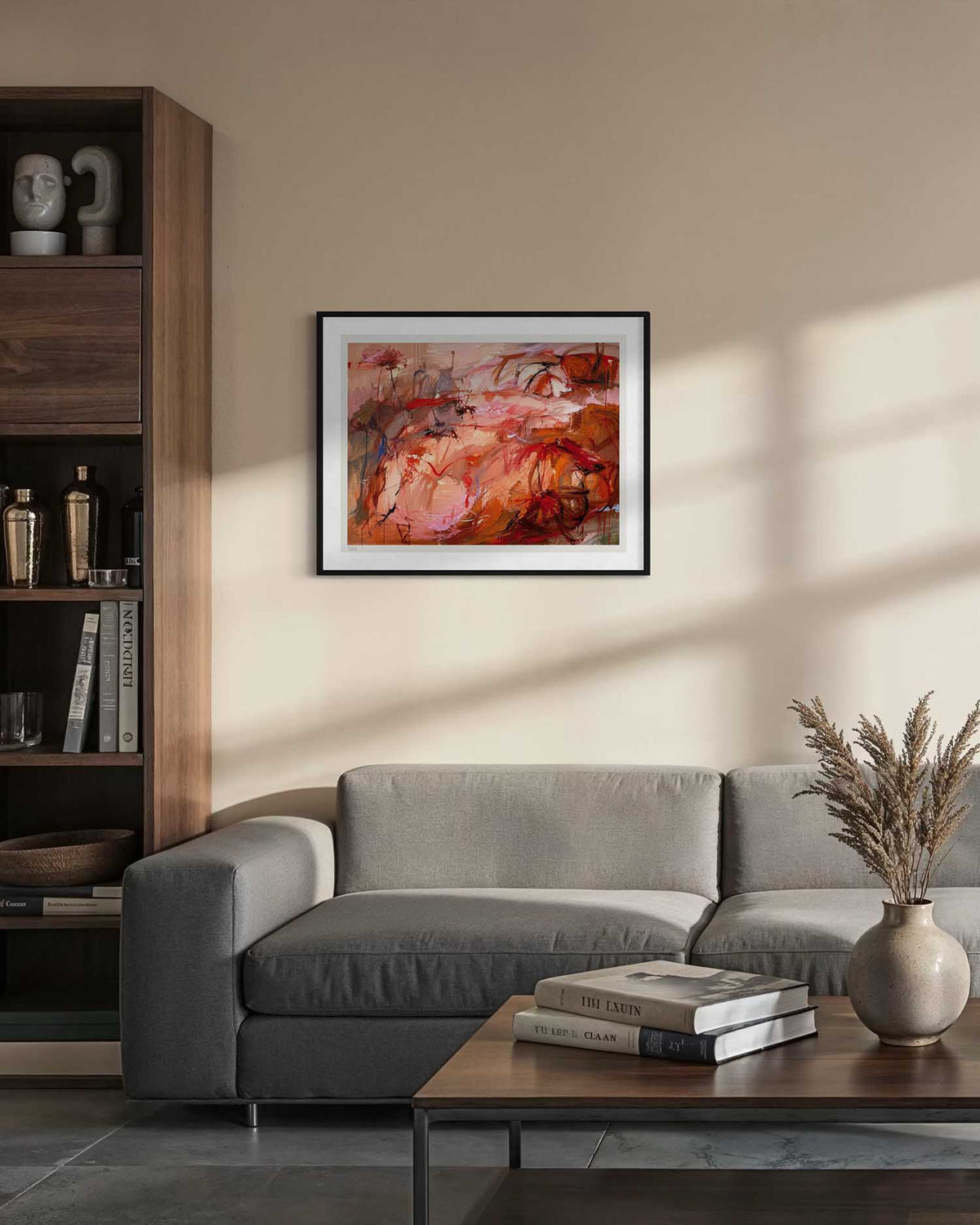 A framed abstract painting with warm red, orange, and pink tones hangs on a beige wall above a gray sofa in a modern living room setting.