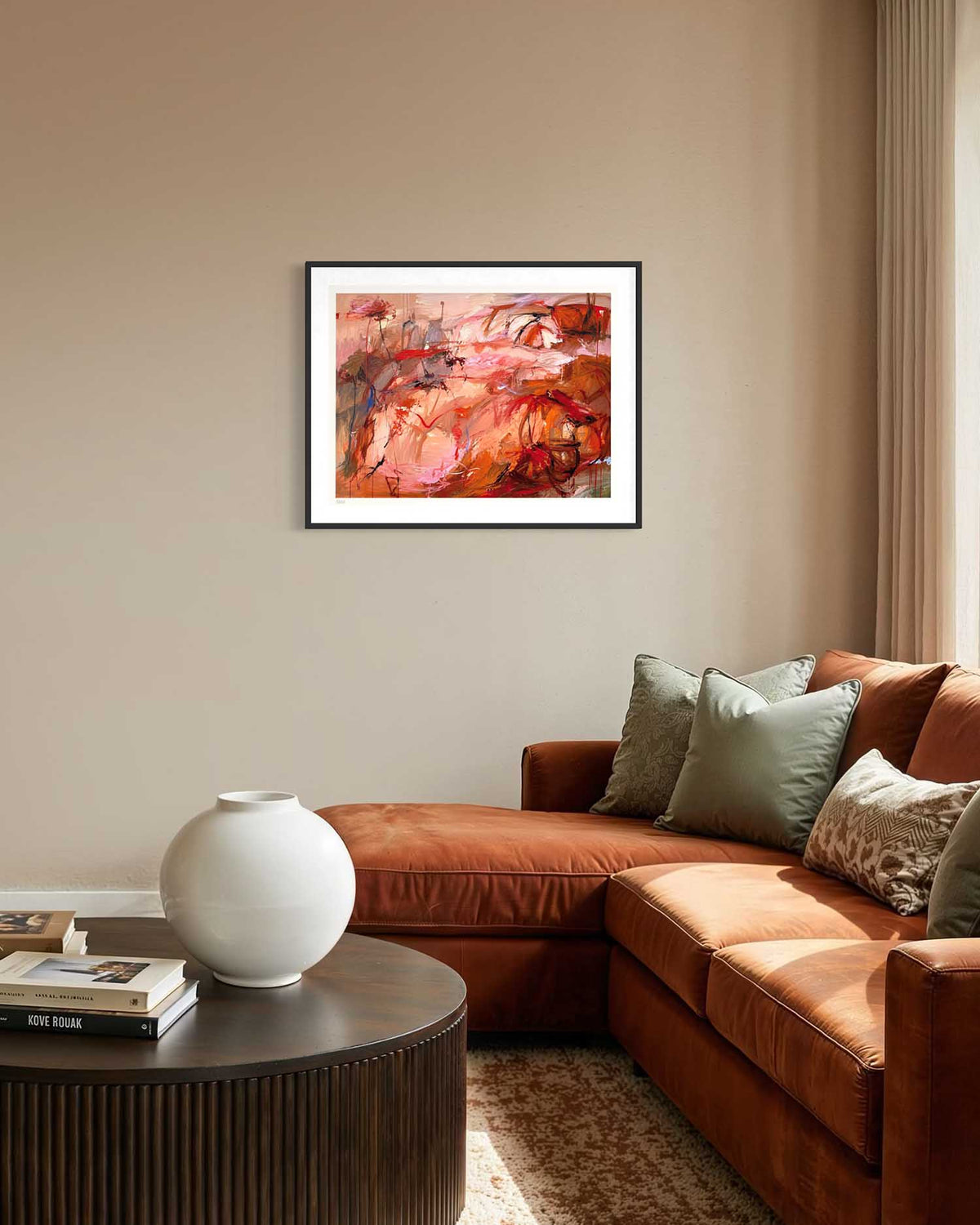 An abstract painting with warm red, orange, and pink tones, displayed in a black frame with a white mat on a beige wall above a modern brown sectional sofa in a living room setting.