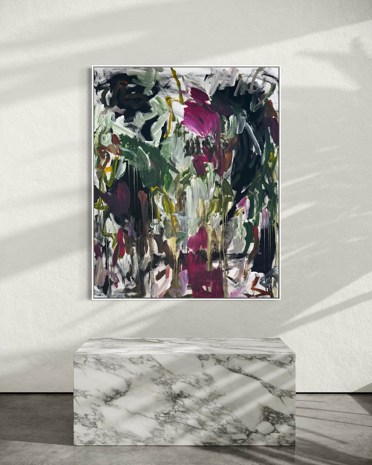 Abstract framed wall art with expressive brushstrokes in green, black, white, gray, and magenta, displayed above a white marble pedestal against a light wall with soft leaf shadows.