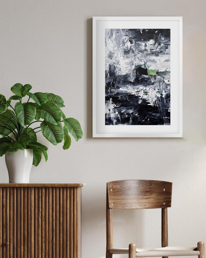 Framed abstract art on a wall above a wooden sideboard with a plant and chair.