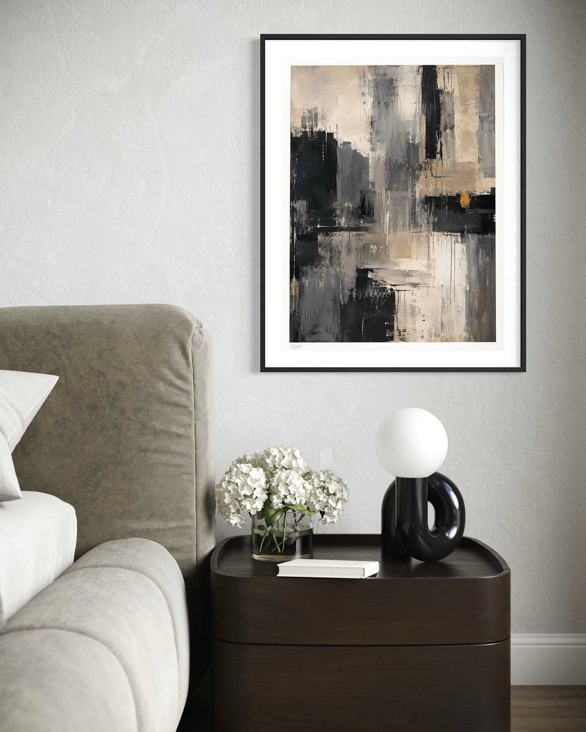 A framed abstract wall art print with black, gray, white, and beige brushstrokes, displayed in a bedroom setting above a dark wood nightstand with a lamp and flowers.