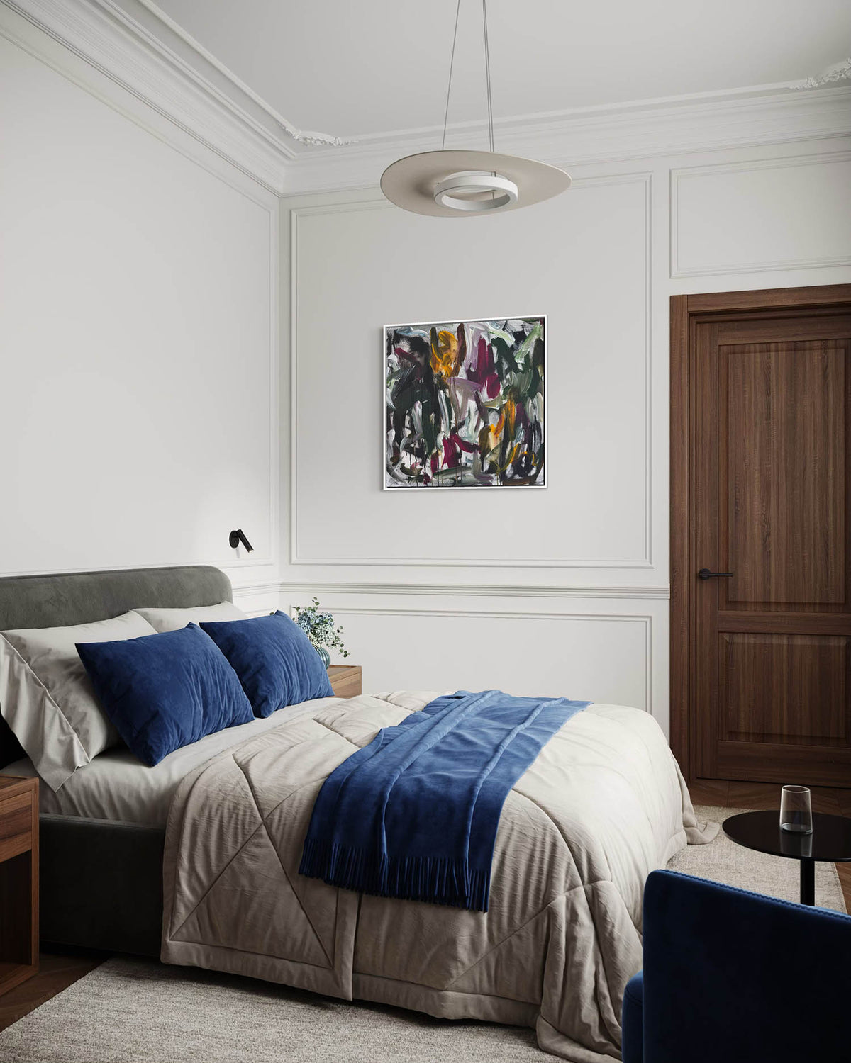 A styled bedroom interior featuring a bed dressed in beige bedding with two dark blue pillows and a blue throw blanket, set against white paneled walls with an abstract painting and a wooden door.