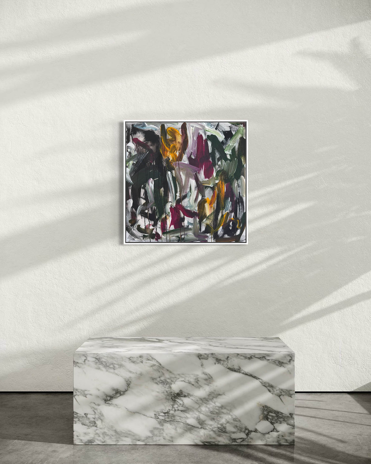 An abstract framed painting with black, gray, white, green, and orange brushstrokes hangs on a textured white wall above a white marble pedestal, with soft leaf shadows cast across the scene.