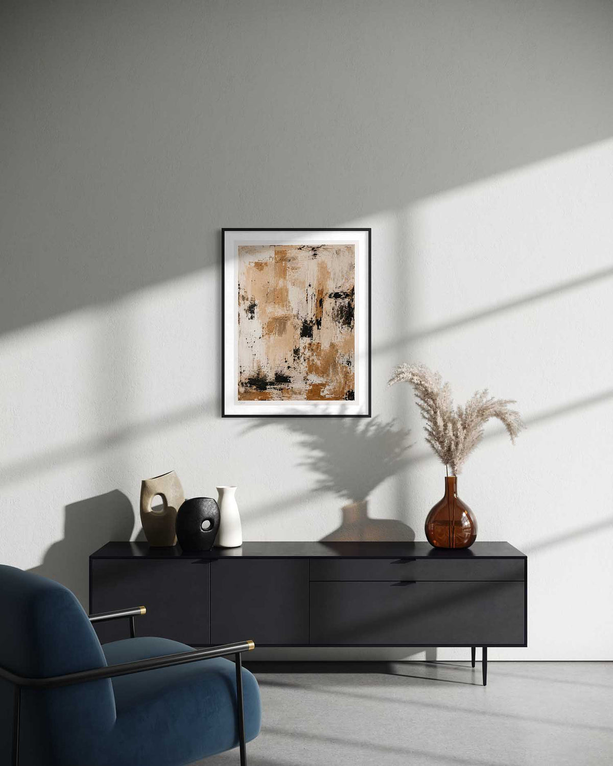 A framed abstract art print with beige, brown, and black tones hangs on a light gray wall above a black sideboard in a modern living room setting with a blue armchair and decorative vases.