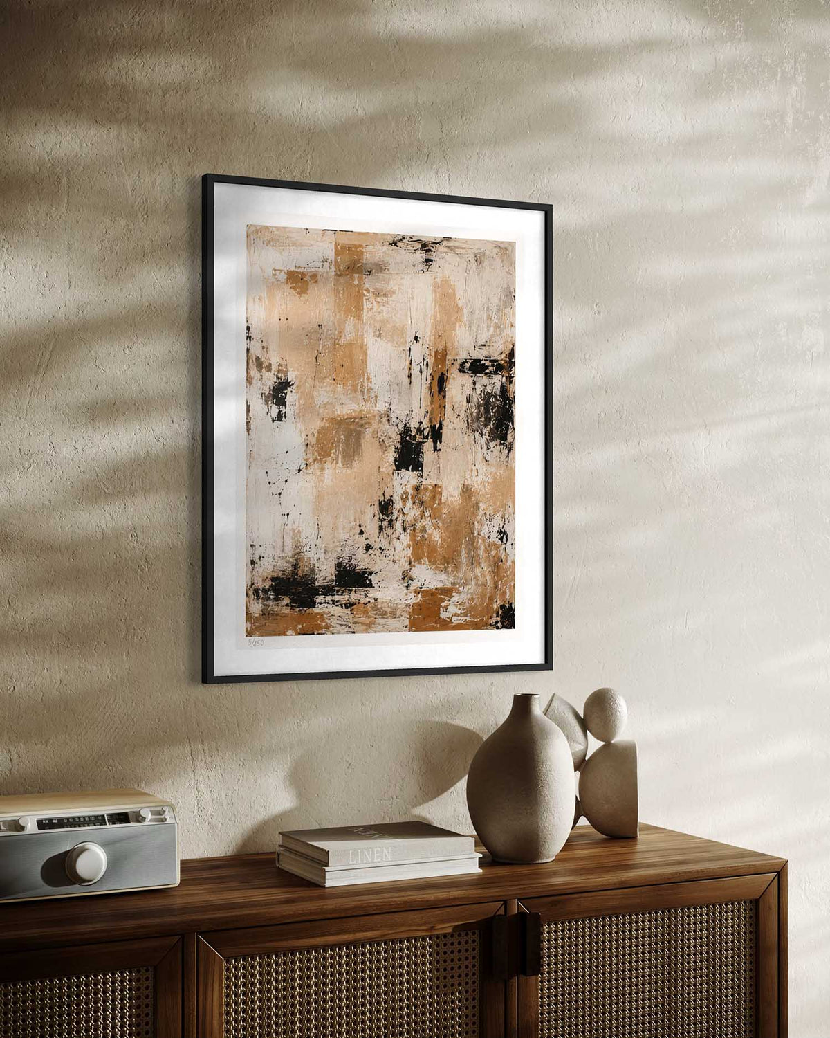 Framed abstract wall art with warm beige, brown, black, and cream tones hanging on a textured beige wall above a wooden cabinet with decorative objects.