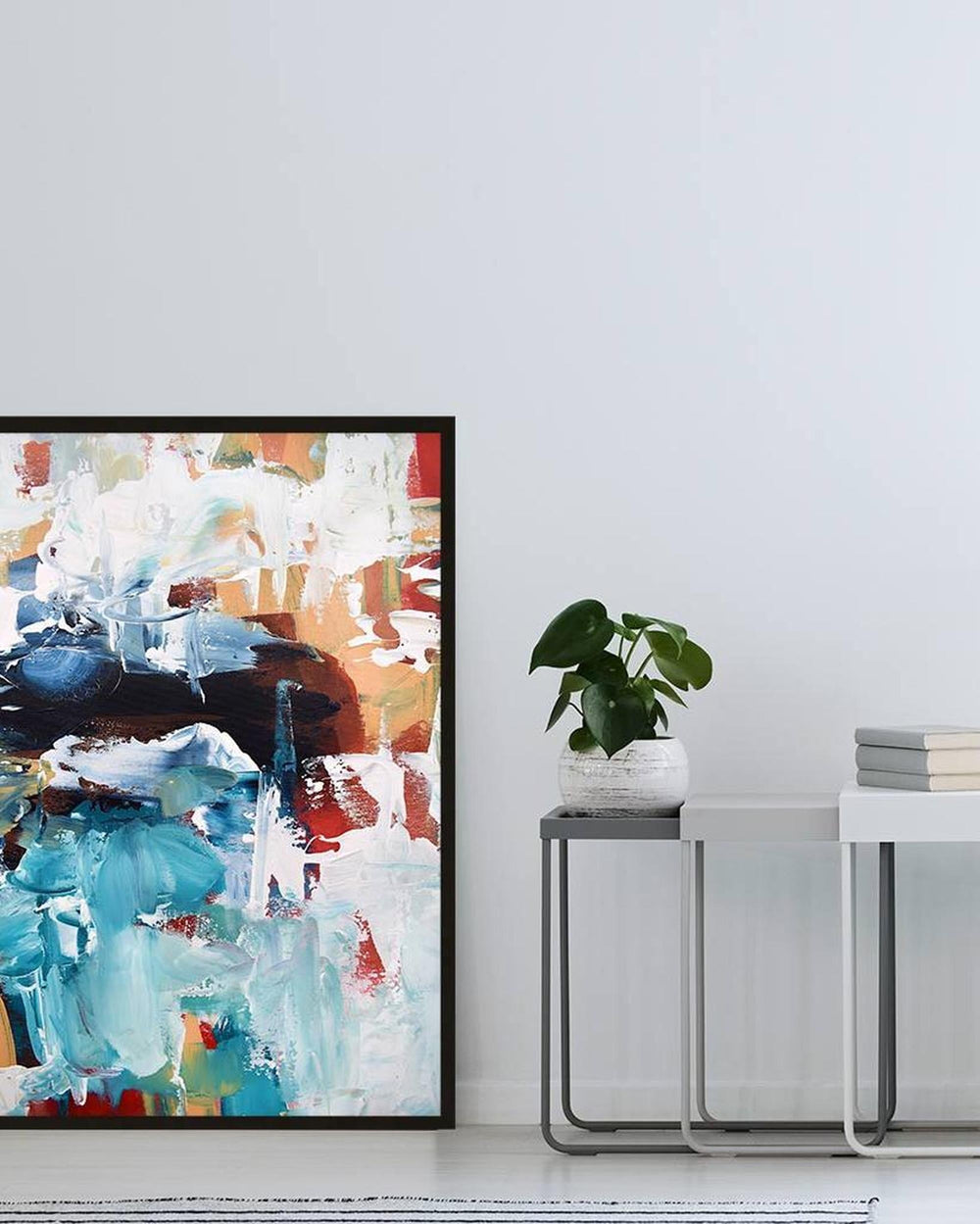 Abstract art print in a black frame on a white wall with a plant and books below.