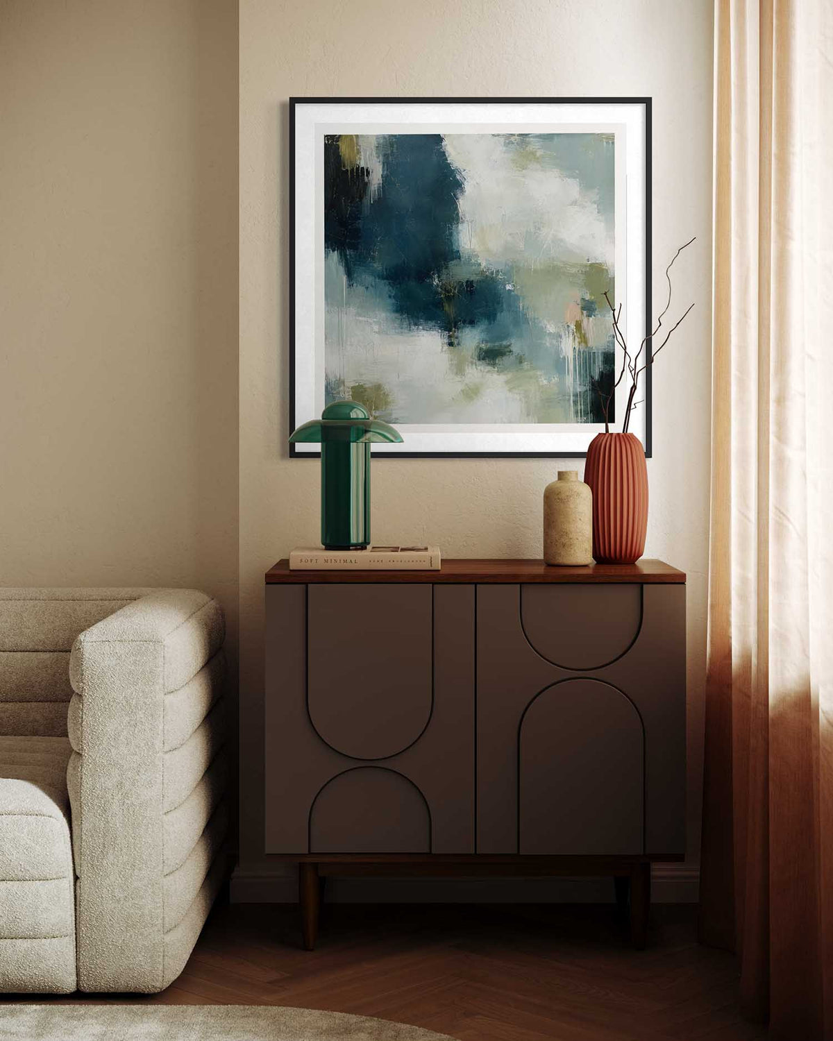 A framed abstract art print with navy, green, and cream tones hangs on a beige wall above a mid-century modern sideboard cabinet in a styled living room setting.