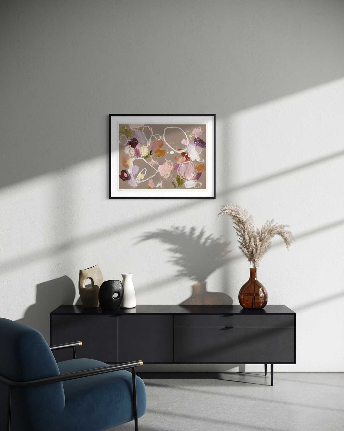 A framed abstract floral art print with pink, purple, and green tones hangs on a light gray wall above a black sideboard in a modern living room setting with a blue armchair and decorative vases.