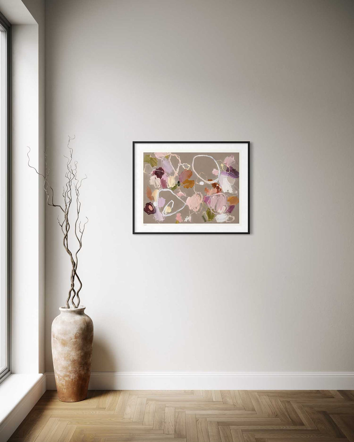 A framed art print depicting a beach landscape with pink sky and green foliage, displayed on a light gray wall in a minimalist room with a wooden herringbone floor and a tall decorative vase.