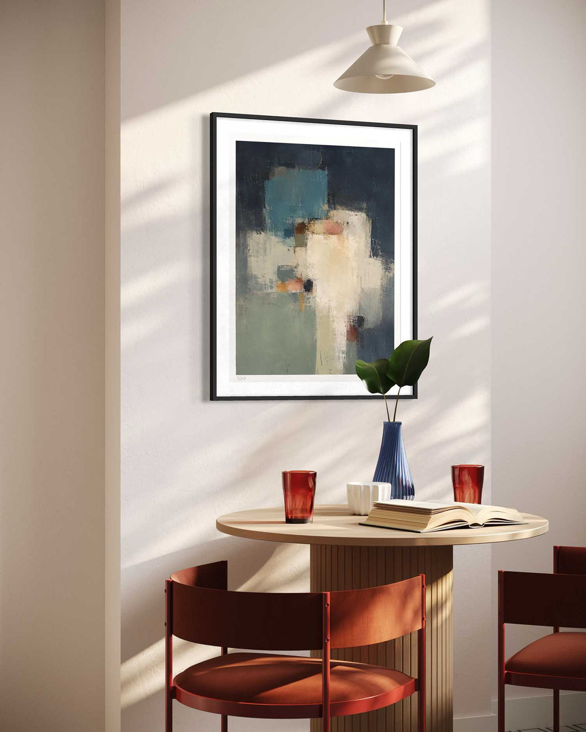 A framed abstract art print hangs on a wall in a modern dining room setting above a round wooden table with red chairs, a blue vase, and a hanging pendant light.