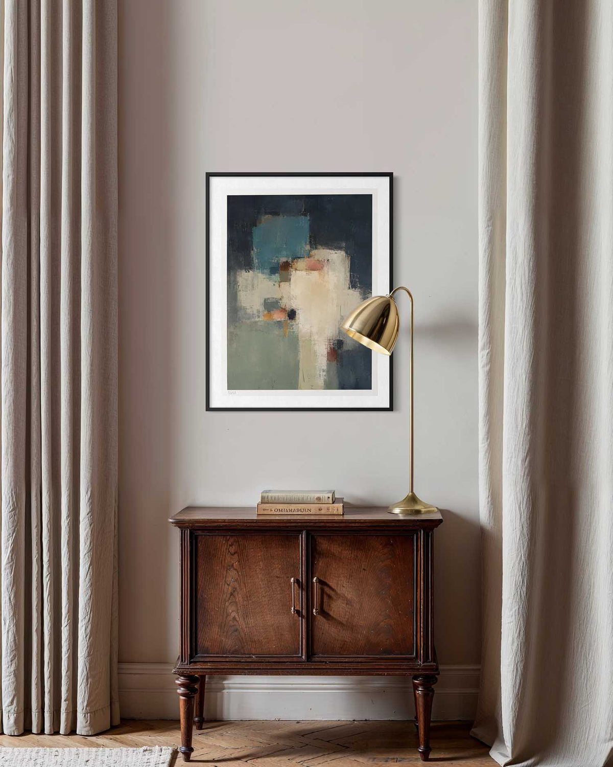An abstract framed wall art print hangs on a beige wall above a dark wood cabinet, flanked by beige curtains and a gold floor lamp.