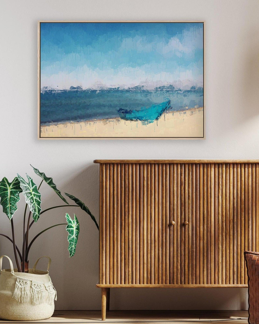 Framed artwork of a blue boat on a beach above a wooden cabinet with a plant and basket below.