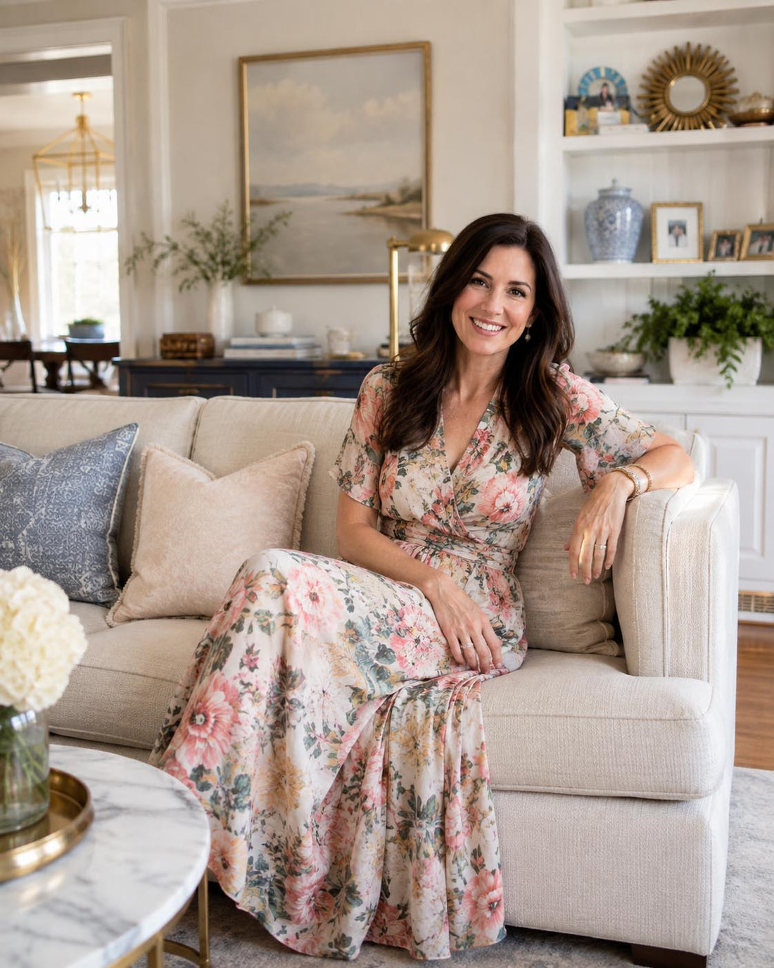 A woman with long dark hair sits smiling on a light beige sofa in a bright, elegant living room. She is wearing a floral wrap maxi dress with short flutter sleeves and a V-neckline in soft pink, peach, and green tones.