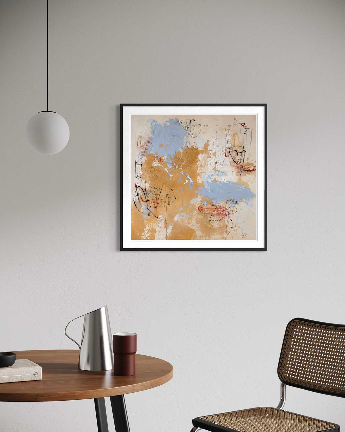 A framed abstract art print with blue, beige, and orange brushstrokes, displayed on a light gray wall in a modern room setting with a wooden table, pendant light, and chair.