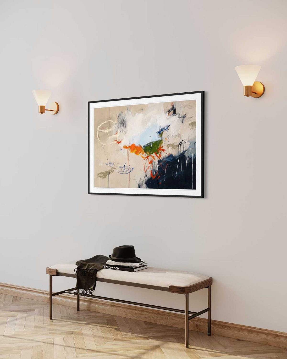 A framed abstract painting with black, white, blue, orange, and green brushstrokes hangs on a light gray wall above a bench with a hat and jacket on it. Two wall sconces light the scene.