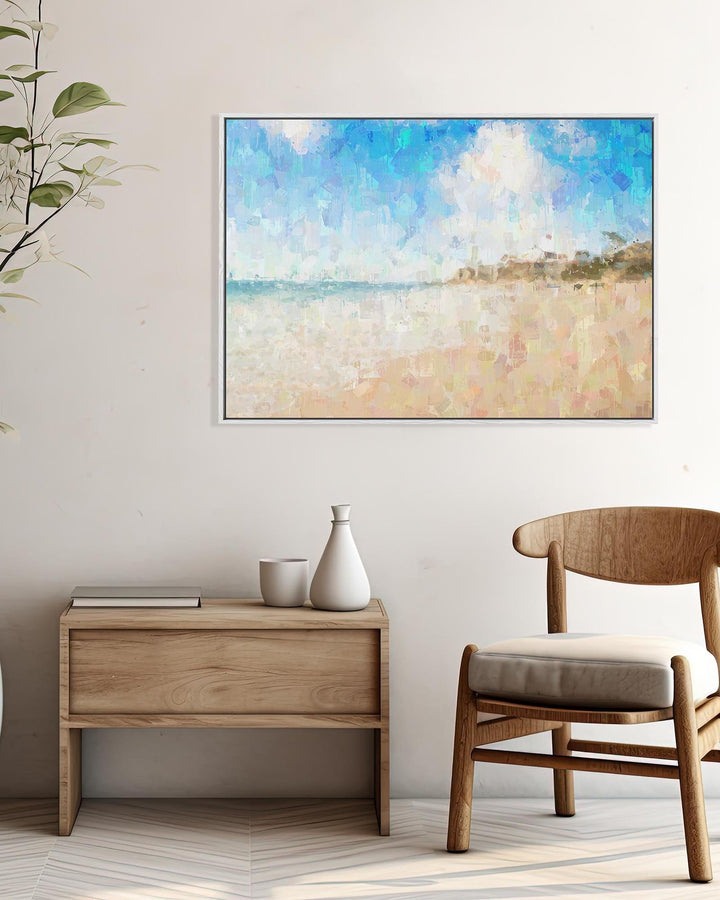 Framed beach artwork on a wall above a wooden side table and chair.