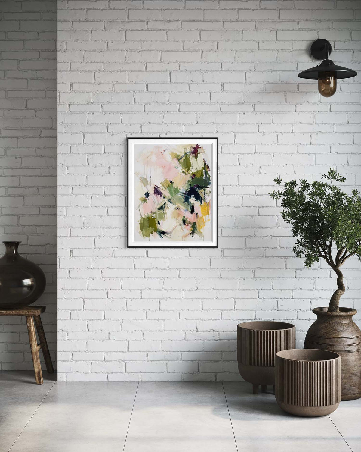 A framed abstract floral art print with soft pink, green, and yellow brushstrokes hangs on a white brick wall in a styled interior setting with a black pendant light, a potted tree, and ceramic vases.