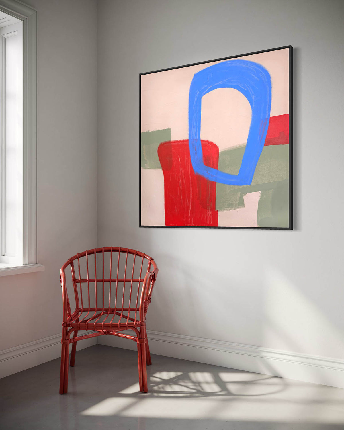 Abstract framed wall art hanging on a light gray wall in a room, featuring a large blue loop shape over red, green, and cream blocks. A red chair sits below the artwork near a window.
