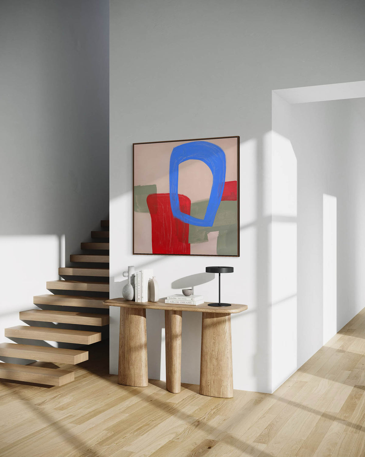 A framed abstract wall art print hangs above a light wood console table in a modern interior with floating stairs, white walls, and warm sunlight casting shadows.