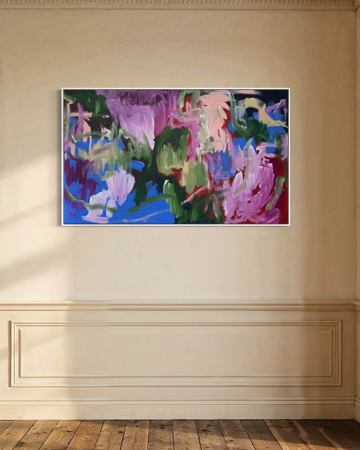 An abstract painting with expressive brushstrokes in purple, pink, blue, green, and black, displayed on a beige wall above a wooden floor in a styled interior setting.