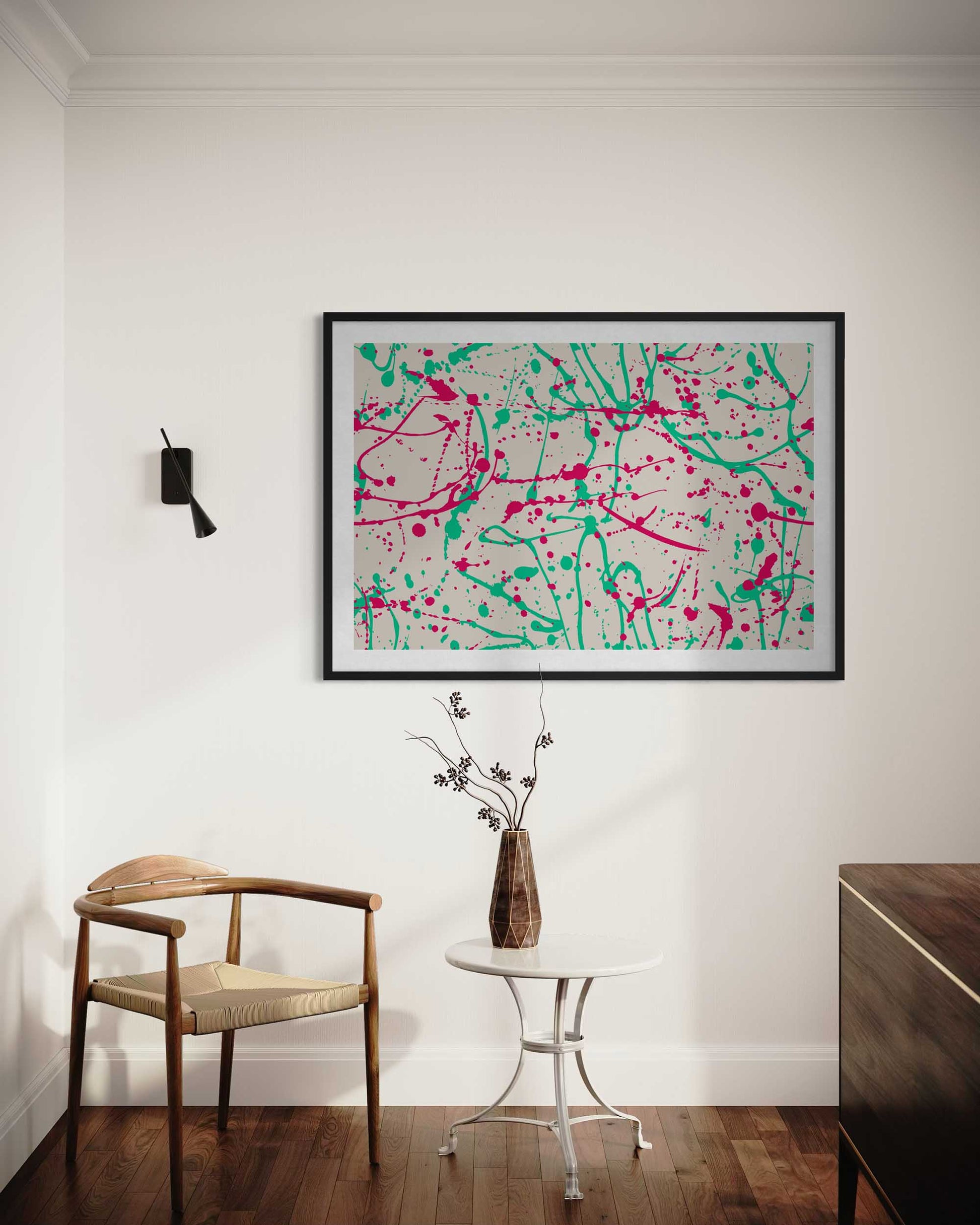 Abstract artwork with pink and green splashes in a black frame on a white wall, with a chair and table in the foreground.