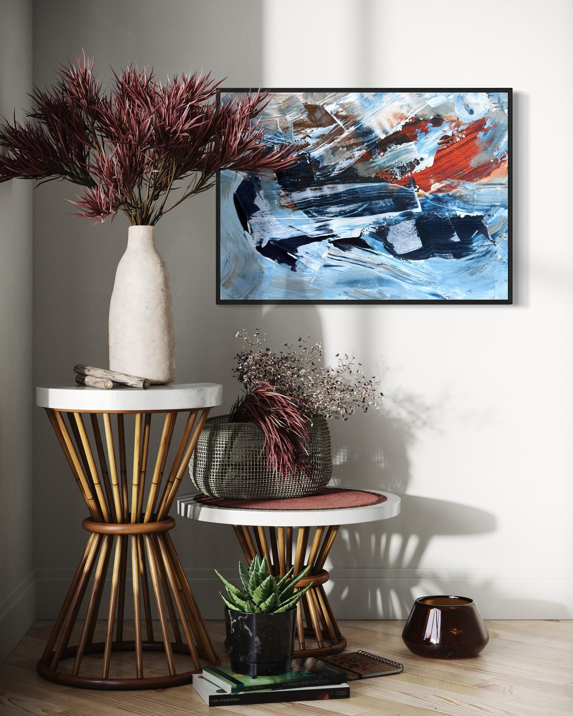 An abstract canvas painting with blue, white, black, and red brushstrokes hanging on a light gray wall in a styled interior setting with side tables, a vase, and dried plants.