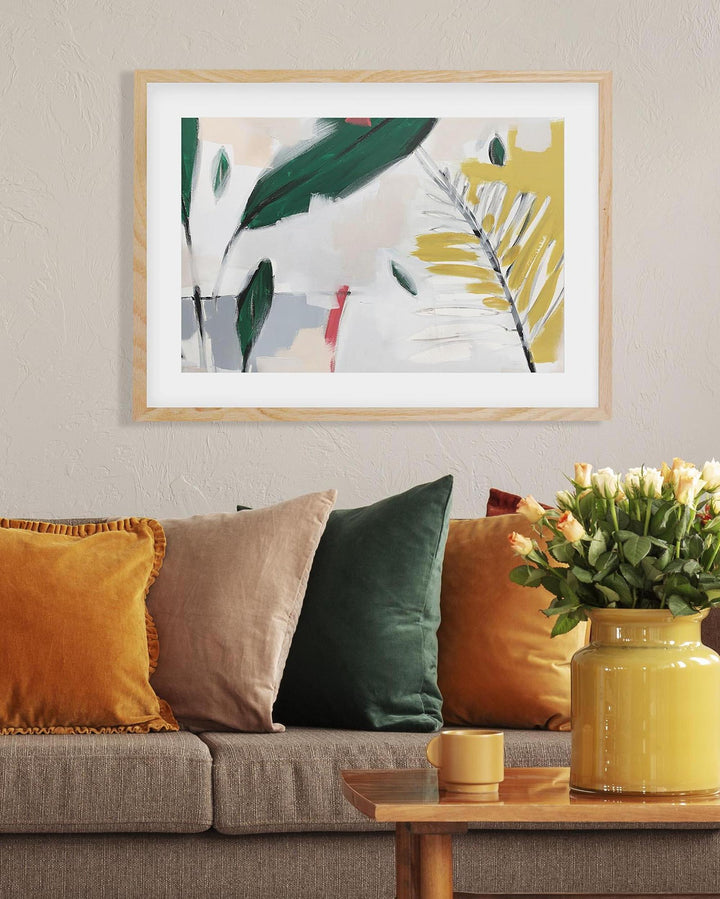 Framed abstract art with leaves above a couch with colorful pillows and a vase of flowers.