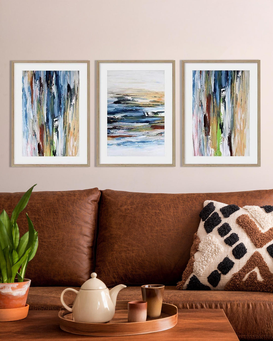 A set of three framed abstract coastal prints displayed on a wall above a brown sofa with a framed picture window让way to the ocean.